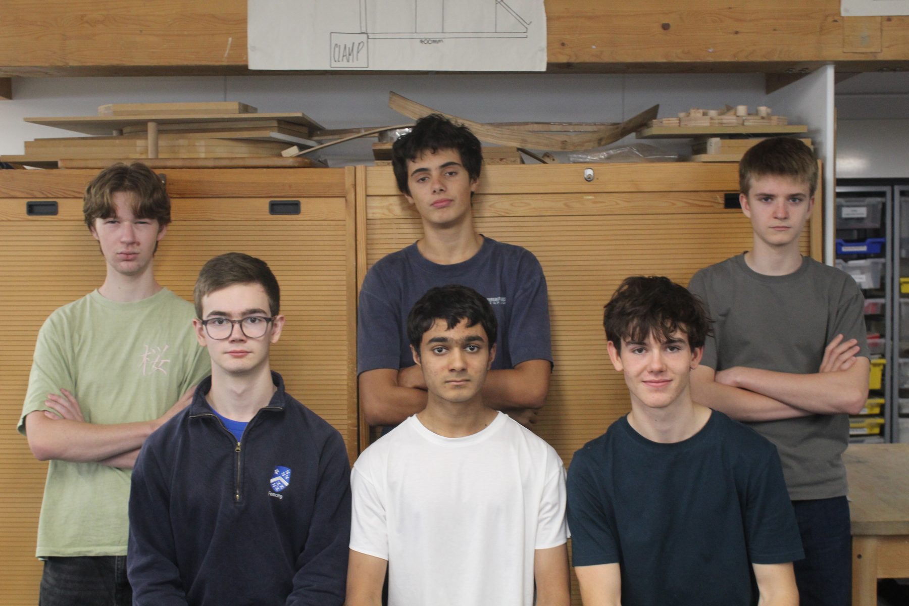 The six members of Swiftly standing with arms folded in their school workshop, in front of timber racking and a pinned technical drawing.